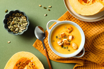 Creamy pumpkin soup with croutons and seeds, styled with raw pumpkin slices. Flat lay, top view.