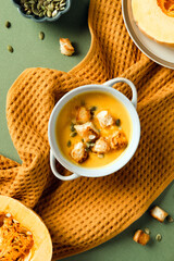 Bowl of vegetarian autumn pumpkin soup with croutons and seeds in a white bowl, surrounded by raw pumpkin, ingredients, and orange towel. Flat lay, top view.