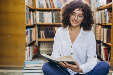 Young student reading a book sitting on library floor