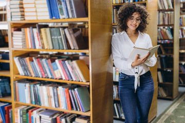 Young student reading a book in the library between bookshelves