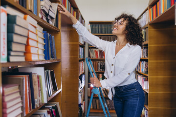 Young librarian picking a book from a high shelf using a ladder