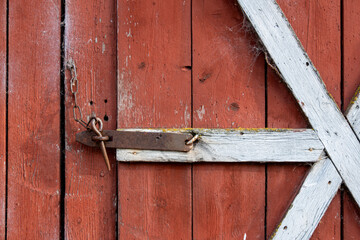 Old rustic latch with a chain securing a weathered red shed door, framed by faded white wooden beams and covered in cobwebs, showing age and rural charm