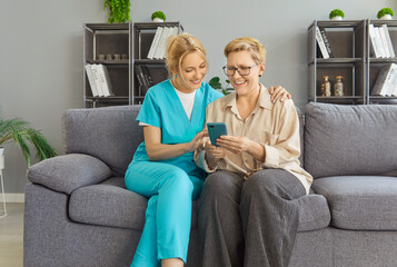 Smiling young woman caregiver sitting on couch beside elderly female in glasses, helping her with...