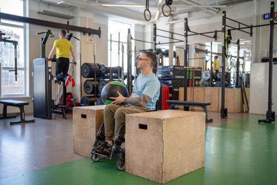 Determined athlete in wheelchair do controlled lifts with weighted ball between parallel bars. Strengthening core muscles post spinal trauma, adaptive sports, neurological recovery, disability fitness