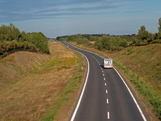 A single-lane asphalt road leads through meadows and forests. The season is summer.