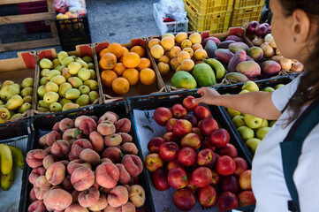 A woman selects vibrant fresh fruits at an outdoor market stall. The array of colorful produce, including peaches, apples, and bananas, highlights healthy food choices and local produce availability.