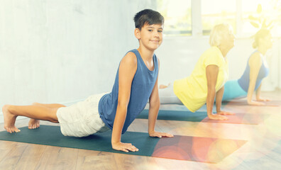Diligent preteen boy practicing upward facing dog pose of yoga in fitness room with other persons