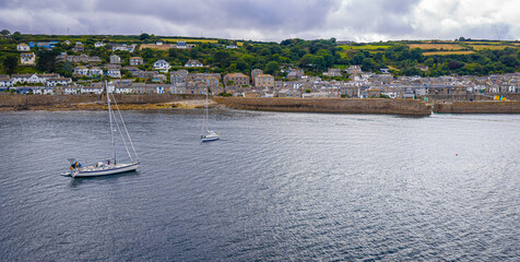 Obraz premium Aerial view of Mousehole harbour in Cornwall, UK, showing moored boats, turquoise waters, sandy shore, and surrounding stone buildings