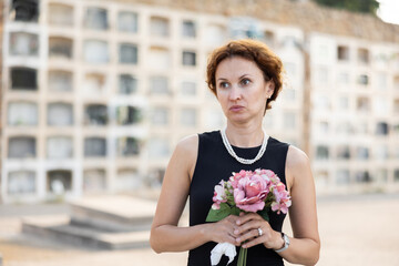 Elegant grieving woman in forties, wearing formal black dress and pearls, standing with bouquet in...