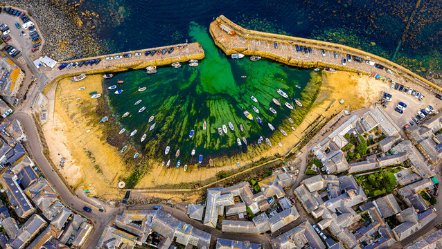 Aerial view of Mousehole harbour in Cornwall, UK, showing moored boats, turquoise waters, sandy shore, and surrounding stone buildings