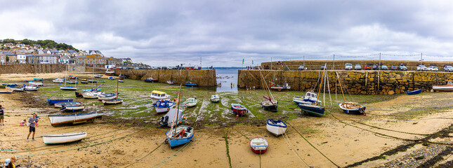 Fishing boats resting on seaweed-covered ground at low tide in Mousehole harbour, Cornwall, with stone pier and parked cars in background