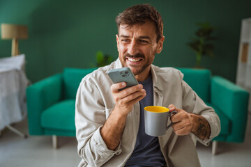 Smiling man using smart phone and drinking coffee at home