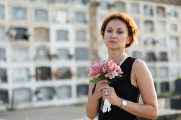 Sorrowful young woman in black dress with bouquet of flowers and handkerchief in hands, stands in...