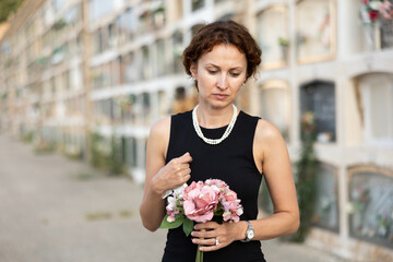 Elegant grieving woman in forties, wearing formal black dress and pearls, standing with bouquet in...