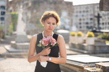 Sorrowful young woman in black dress with bouquet of flowers and handkerchief in hands, stands in...