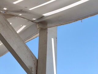 White wooden pergola beams and posts against a clear blue sky, casting shadows on a canvas roof....