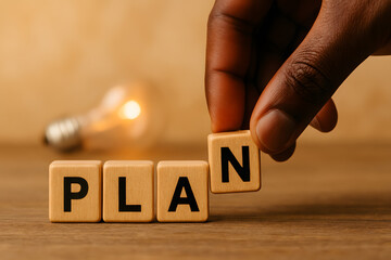 Strategic Plan Concept: The close-up view emphasizes a hand carefully arranging letter tiles, spelling out 'PLAN', underscored by a softly glowing light bulb in the background.