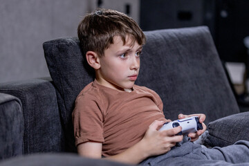 Boy is sitting on sofa at home and playing video games with his joypad.