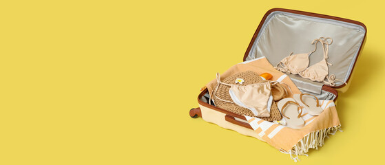 Open suitcase with stylish swimsuit and beach accessories on yellow background, closeup