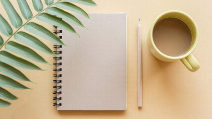 Workspace flatlay featuring spiral notebook, wooden pencil, coffee mug and palm leaf on light yellow background