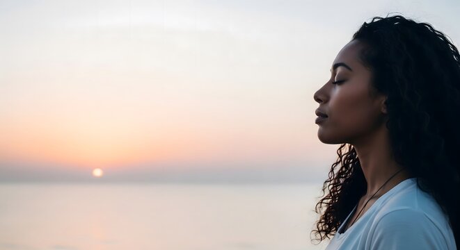 Profile of a serene Black woman with long curly hair and closed eyes, meditating or relaxing on a beach during a peaceful sunset over the ocean.