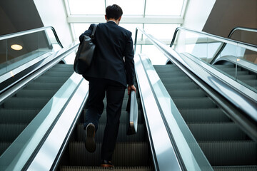 Businessman seen from behind, carrying a briefcase while riding up an escalator in an airport or modern office building, symbolizing career progression and moving up
