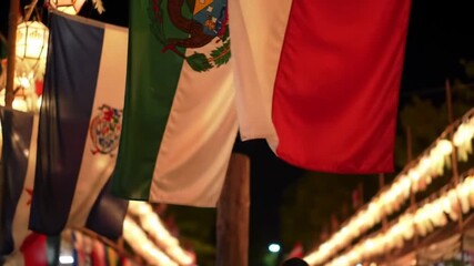 National flags of El Salvador, Mexico, and Italy hanging illuminated at night during an outdoor cultural celebration with string lights.