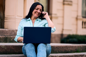 Beautiful mature Latina working on her computer in her backyard, smiling happily, while also...