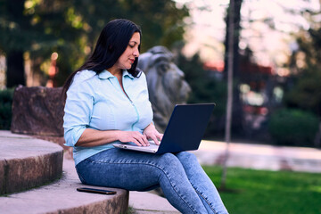 Beautiful mature Latina woman working on her laptop outdoors at home, smiling, while also using her...