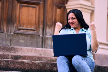 Beautiful mature Latina woman working on her laptop outdoors at home, smiling, energetic, and very...