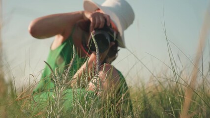 woman wearing white hat and green dress kneeling in grassy field using camera to photograph wild plant during sunny outdoor session with soft natural light and visible focus on subject in foreground