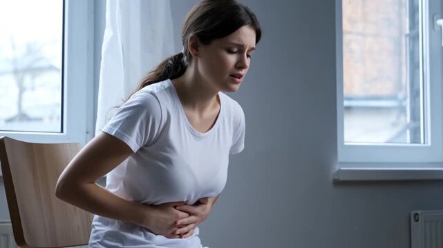 A distressed woman sits, clutching her stomach and wincing in pain, her body language a vivid portrayal of discomfort and a silent battle with internal physical distress.