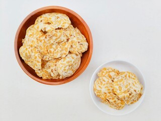 Tempeh chips in a white plate and a wooden plate on a white background. 