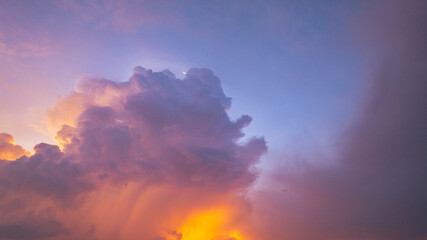 A vivid and dynamic sky captures towering storm clouds illuminated by the warm hues of a setting sun, creating a striking contrast between light and shadow above a darkened forested landscape.