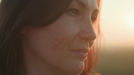 Close up of young woman gazing downward with calm expression as golden sun sets behind her, soft wind moves hair gently across her face in tranquil outdoor atmosphere filled