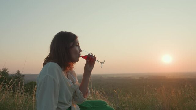 Woman seated on grassy hillside sipping red wine from glass as sun sets over forest horizon, soft light casting glow across sky and landscape creating peaceful evening mood with fruit on ground