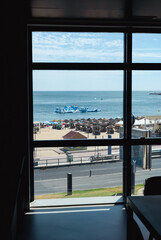 View of Santo Amaro beach in Oeiras, Portugal, seen through a large window. The scene shows the sandy beach with sun umbrellas, people enjoying the seaside, and a floating water park structure