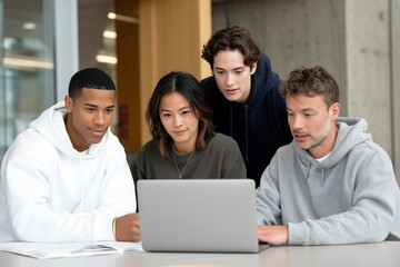 Diverse group of students collaborating and studying together around laptop computer in modern classroom or campus workspace