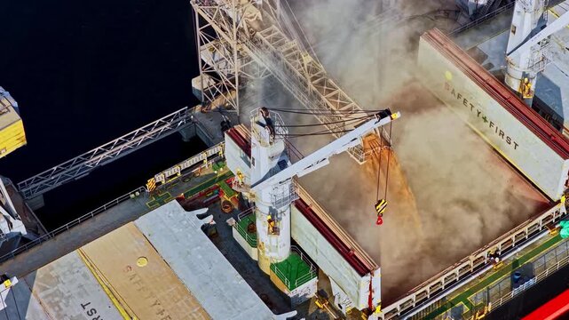 A dynamic top-down aerial parallax shot shows a crane and chute loading grain into the open hold of a large bulk carrier ship at the industrial Port of Riga, Latvia.