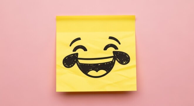 A yellow sticky note with a hand drawn laughing emoji on a pink background in a studio setting light