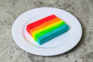 Close up view of one rainbow cake on the white plate. Grey or gray Cement. Background. colourful. Colorful. 