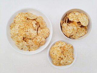 Tempeh chips in a white plate and a small white plate and a white bowl on a white background. Top of view. 