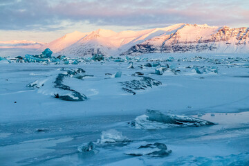 Icebergs and huge chunks for green-blueish ice fill Jokulsarlon Glacier lagoon, as the golden hour sunlight of the early morning casts a beautiful warm glow over the glacier-filled national park in