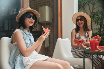 Chilling Asian family holidays, casual girl and mum in sunglasses enjoy eating fresh watermelons sliced and smoothie drinks, happiness relaxing on vacation, and hot summertime at outdoor terrace.