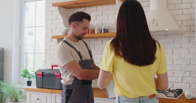 Smiling handyman in uniform shaking hands with woman client in kitchen after repair service. Young woman thanking professional repairman, technician or plumber after completing home maintenance.