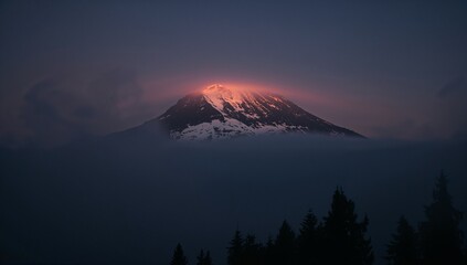 Fototapeta premium Snowy mountain peak glowing at sunrise above the clouds and trees