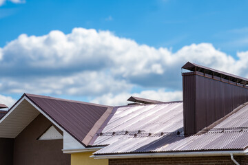 Fototapeta premium The roof of a three-storey house made of brown metal tiles against a blue sky background. There is ventilation on the roof