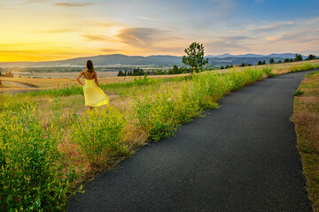 A young woman in a yellow dress stands near a walking trail overlooking the Spokane Valley at sunset in Washington State at summer.