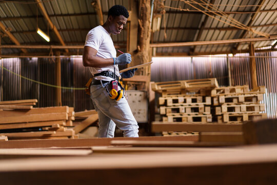 Black carpenter working in woodworking factory, carefully inspecting a wooden plank after cutting. Concept of craftsmanship, labor, and skilled profession.