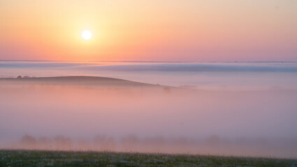 Serene sunrise over a misty valley with rolling hills bathed in soft pastel light and a gentle haze creating a dreamlike atmosphere